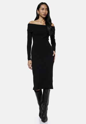 Vrouw met lang zwart haar die een nauwsluitende zwarte off-shoulder jurk en zwarte leren kniehoge laarzen draagt, staand tegen een egale achtergrond.