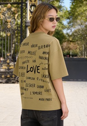 Young woman wearing sunglasses and an olive-green t-shirt printed with the word "love" in multiple languages, standing outdoors near ornate gates.