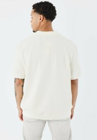 Chemise courte beige clair à manches courtes avec un tissu texturé, col et poignets côtelés. Vue arrière montrant un motif subtil et une coupe décontractée.