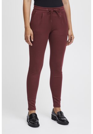 Burgunderfarbene Jogginghose mit einem elastischen Bund und verstellbarem Kordelzug. Slim-Fit, weicher Stoff und seitliche Taschen. Kombiniert mit schwarzen Slippern.