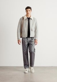 Tânăr îmbrăcat cu o jachetă texturată deschisă la culoare, cu guler alb, cămașă neagră, pantaloni gri, adidași albi și purtând un rucsac bej.