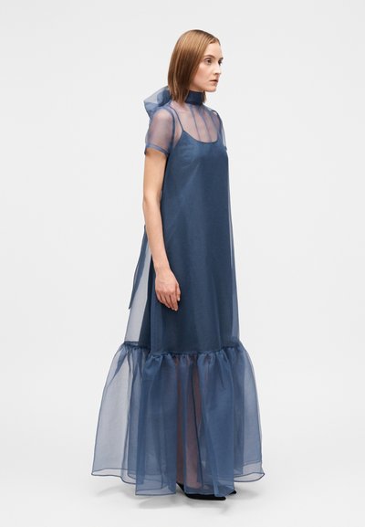 Γυναίκα που φοράει ένα διάφανο φόρεμα σε σκούρο μπλε χρώμα μέχρι το πάτωμα, με κοντά μανίκια, ψηλό λαιμό και βολάν στο στρίφωμα, στέκεται μπροστά σε ένα λευκό φόντο.