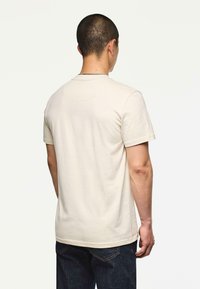 Mężczyzna stojący tyłem, ubrany w gładką, jasnobeżową koszulkę z krótkim rękawem i ciemnoniebieskie dżinsy, na jednolitym białym tle.