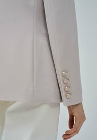Blazer grigio chiaro con texture diagonale. I notevoli bottoni dorati sulle maniche aggiungono dettagli. Forma aderente, tessuto liscio, design fitted.