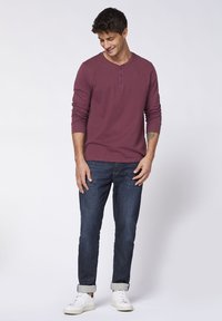 Bordeauxfarbener Langarm-Henley-Shirt mit Drei-Knopf-Leiste, kombiniert mit dunkelblauen Jeans und weißen Sneakers, mit aufgerollten Ärmeln.