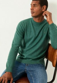 Grüner gestrickter Pullover mit Rundhalsausschnitt und gerippten Bündchen, kombiniert mit blauen Jeans. Der Pullover hat ein dezentes Logo auf der Brust.