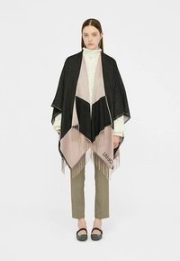 Schwarzer und beiger Poncho mit geometrischem Design, mit Fransen an den Rändern und einem Markenetikett. Glatte Textur, leichtes Material.