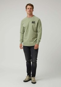Hombre en sudadera verde con el texto "DREAM BIG", jeans negros y zapatillas beige, de pie contra un fondo claro y liso.