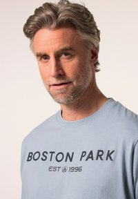 Himmelblaues Baumwoll-T-Shirt mit "BOSTON PARK EST 1996" in dunkler, gestickter Schrift. Rundhalsausschnitt und kurze Ärmel. Glatte Textur.