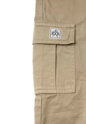Beige stof cargolomme med klap og påsyet hvid lap med krydsede bogstaver B og R med nummer 36 over dem.