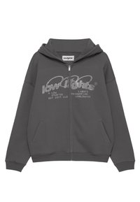 Sudadera gris con cremallera que presenta una capucha con cordón y bolsillos laterales. La parte frontal muestra un logotipo gráfico texturizado en plata. Fabricada con tela suave.