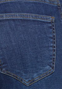 Yksityiskohta sinisestä farkkukankaasta, jossa on ommeltu takatasku, jossa on kultaruskeat langat.