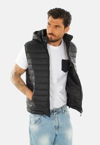 Gilet gonfiabile nero con cappuccio, dotato di una chiusura lampo sul davanti, tasche laterali e un design trapuntato. Indossato sopra una maglietta bianca.