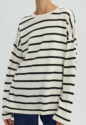 Personne portant une chemise blanche ample à manches longues avec des rayures horizontales noires et un pantalon bleu foncé, cadré au niveau du cou et des hanches.