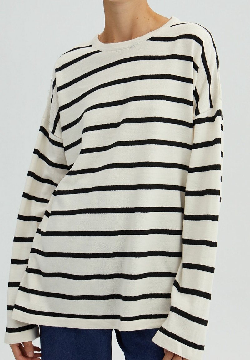 Personne portant une chemise blanche ample à manches longues avec des rayures horizontales noires et un pantalon bleu foncé, cadré au niveau du cou et des hanches.