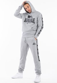 Gray cotton hoodie with logo design, matched with gray pants featuring side stripes. White sneakers complete the athletic look.