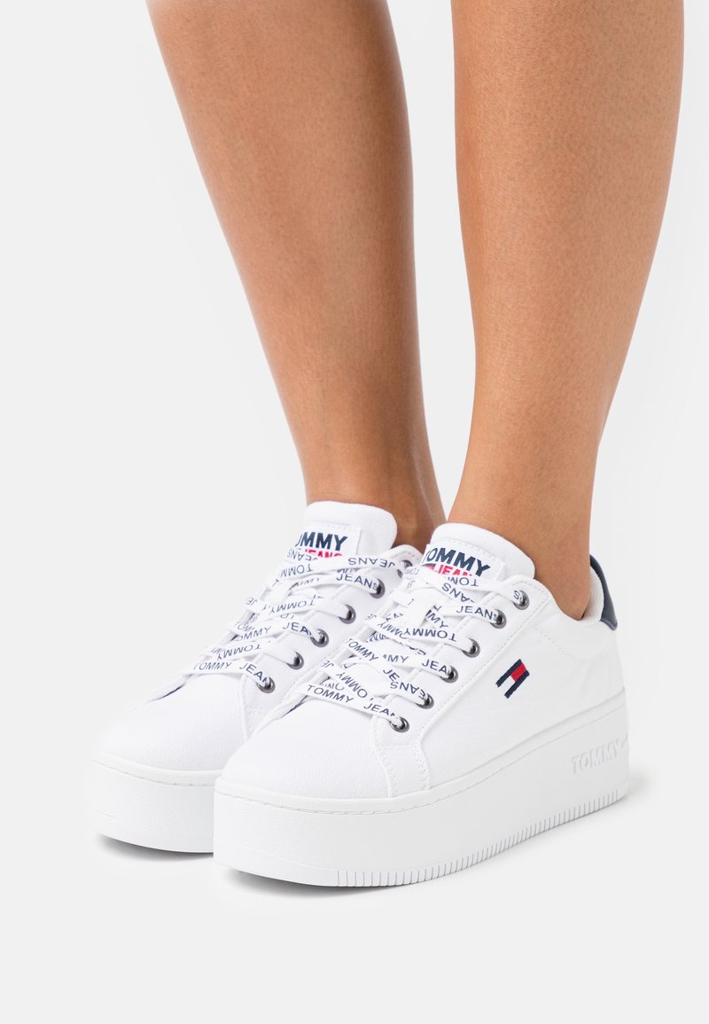 Baskets en toile blanches avec une semelle épaisse, arborant des accents de logo en noir et rouge et des lacets décoratifs imprimés avec "TOMMY JEANS".
