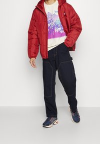 Chaqueta acolchada roja con capucha sobre un suéter claro con un gráfico colorido de montañas, combinada con pantalones anchos azul marino y zapatillas deportivas.