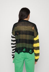 Pull en maille à coupe ample avec des rayures noires, vertes et jaunes, et un ourlet festonné. Le dos présente des ouvertures usées et un détail en dentelle.