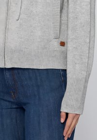 Szary sweter robiony na drutach o ribbowanej fakturze, z przednimi kieszeniami oraz małą brązową skórzaną metką. Połączony z ciemnoniebieskimi dżinsami.