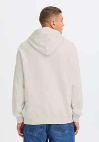 En person med kort hår iført en lysegrå hættejakke, der vender væk, parret med blå jeans, mod en simpel hvid baggrund.