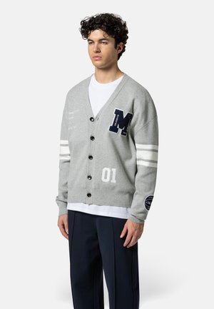 Junger Mann trägt eine graue, geknöpfte Strickjacke mit weißen Streifen an den Ärmeln, einem marineblauen "M"-Aufnäher und der Nummer "01", kombiniert mit schwarzen Hosen und weißem Hemd.