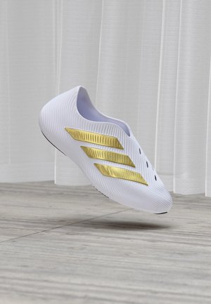 Fehér slip-on sportcipő texturált felülettel és három arany színű átlós csíkkal, lebegve bemutatva egy csempézett padló felett, hátterében áttetsző függönyökkel.