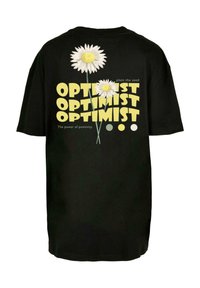 Černé tričko s bílými a žlutými sedmikráskami. Text zní "OPTIMIST" tučně, v opakujícím se vzoru s frázemi "zasadit semeno" a "síla pozitivního myšlení."
