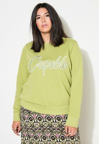Felpa verde con la scritta "Capable" in bianco corsivo. Presenta un colletto rotondo e maniche lunghe. Indossata con una gonna a fantasia.