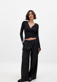 Schwarzes Wickeloberteil mit langen Ärmeln, tiefem V-Ausschnitt und gebundenem Detail. Kombiniert mit schwarzen weit geschnittenen Hosen und strukturierten schwarzen Schuhen.
