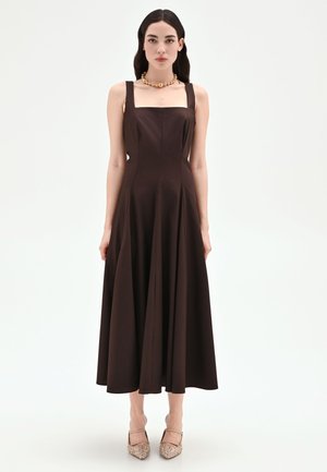 Donna con capelli lunghi e scuri, che indossa un vestito midi marrone scuro senza maniche e tacchi beige, in piedi davanti a uno sfondo chiaro e uniforme.