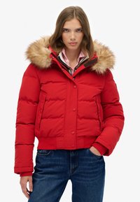 Veste rembourrée rouge avec capuche en fourrure, fermeture éclair et boutons-pression, poches latérales et poignets côtelés, associée à un jean en denim.