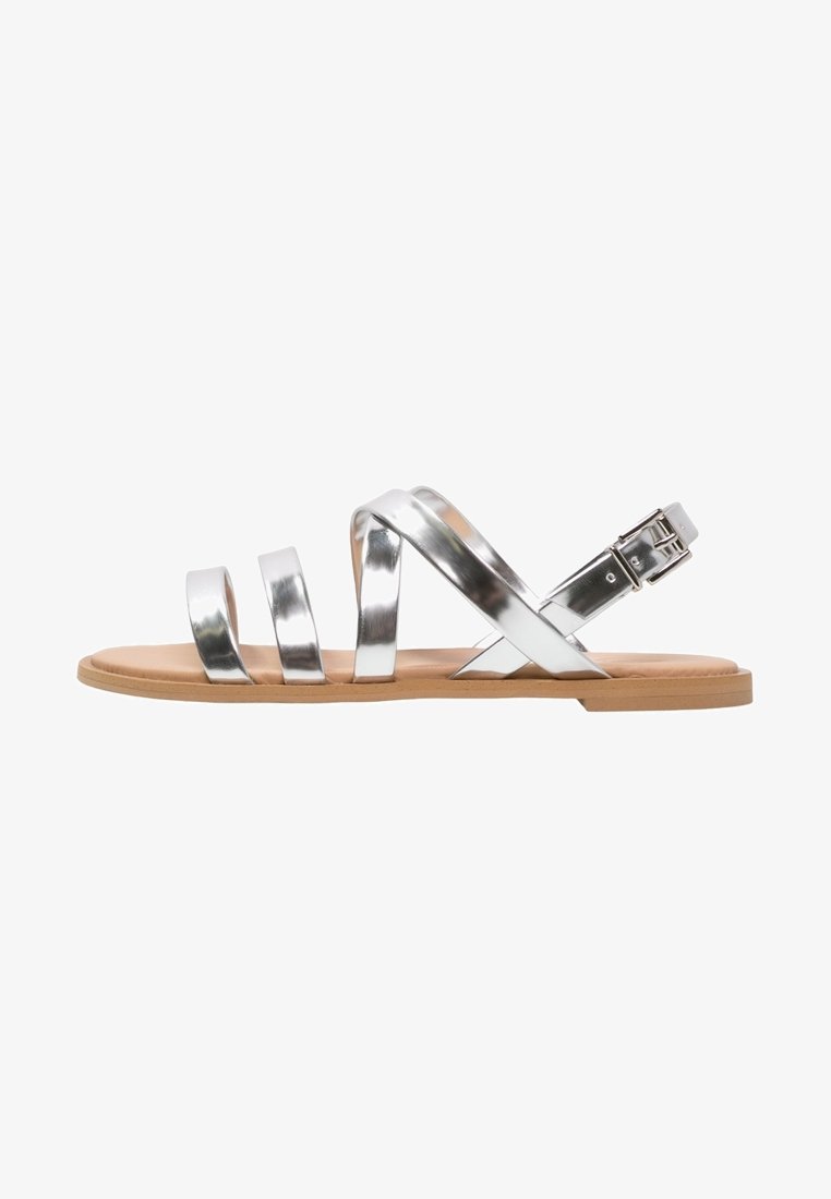 Silberne, metallische Riemchensandalen mit flacher, brauner Sohle, die drei breite Riemen und einen Schnallenverschluss für eine verstellbare Passform aufweisen.