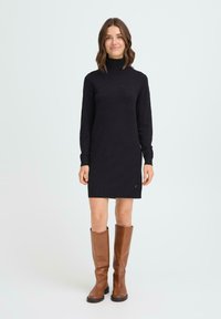 Vestido de punto negro con cuello alto y mangas largas, alcanzando a mitad del muslo. Combinado con botas de cuero marrón hasta la rodilla. Detalle de pequeño logo en el dobladillo.