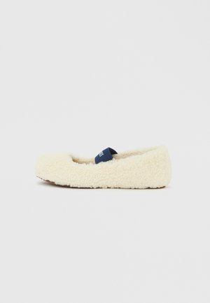 Pantofole color crema con una fascia blu navy sopra, viste di lato su uno sfondo bianco semplice.