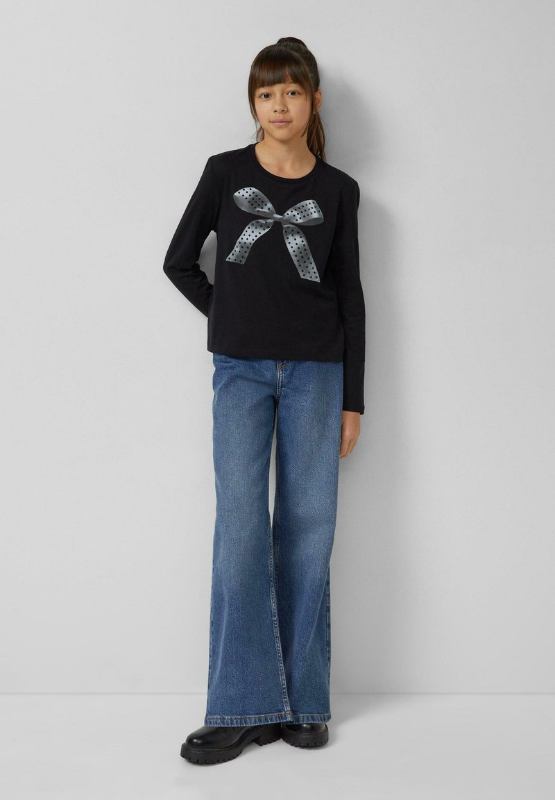 Schwarzes Langarmhemd mit grauem Polka-Dot-Schleifengrafik, kombiniert mit weit geschnittenen blauen Jeans und schwarzen klobigen Schuhen.