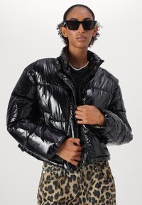 Černá puffa bunda s lesklým povrchem, polstrovaným designem a patentkami, nošena přes černý top a džíny s leopardím potiskem.