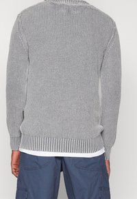 Harmaa neulepaita, jossa on resorit hihansuuissa ja helmassa. Siinä on teksturoitu kuvio, pyöreä pääntie ja se on kerrospukeutunut valkoisen paidan päälle.