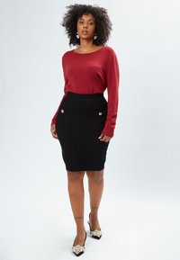 Maglione rosso a maniche lunghe, gonna a tubino nera aderente con texture trapuntata e dettagli in oro, abbinata a tacchi neri decorati con perle.