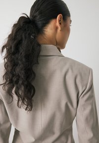 Ljusgrå blazer med skarpa axlar och skräddarsydd passform. Texturerad tyg synlig på baksidan, med en notch-krage och rena linjer.