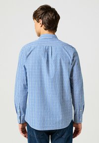 Camisa a cuadros azul y blanca de algodón, con mangas largas, cuello abotonado y espalda ventilada para mayor comodidad al moverse.