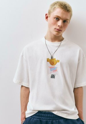 Jeune personne aux cheveux blonds courts portant un T-shirt blanc oversize avec un motif d'ourson en gomme et un pantalon bleu, debout contre un mur blanc.
