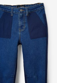 Vaqueros de denim con una tela de color azul oscuro, que presentan secciones de azul más claro, dos grandes bolsillos frontales y costuras naranjas visibles a lo largo de las costuras.