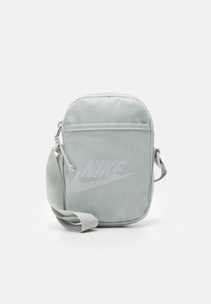 Petit sac bandoulière gris clair avec poche avant en maille et sangle réglable, arborant un logo Nike swoosh discret sur la poche.