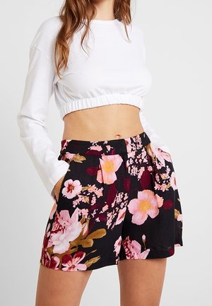 Woman wearing a white long-sleeve cropped top and high-waisted black shorts with a pink and burgundy floral pattern.