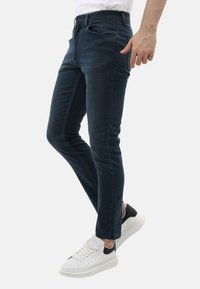 Donkerblauwe denim jeans met een slim fit, voorzien van voorzakken en een knoopsluiting, gecombineerd met witte sneakers en een zwart accent.