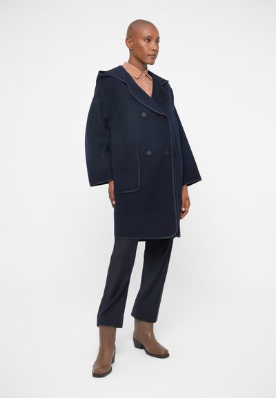 Palton din lână bleumarin, cu două rânduri de nasturi, cu glugă mare, buzunare laterale și detalii contrastante. Asortat cu pantaloni închisi la culoare și cizme maro până la gleznă.