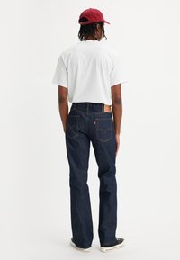 Person iført en hvid t-shirt, mørkeblå jeans, sorte sneakers og en rød kasket, set bagfra mod en ensfarvet hvid baggrund.