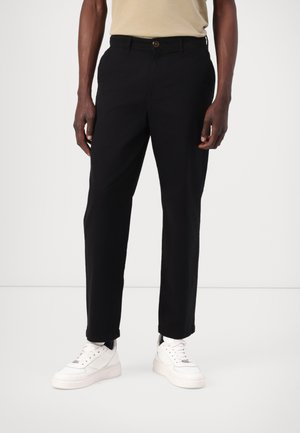 Pantalon en coton noir avec une coupe droite, doté d'une fermeture à bouton et de poches latérales, associé à des baskets blanches.