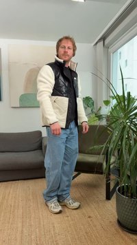 Black and cream fleece jacket, blue jeans, and light sneakers. Background includes a gray sofa, green plant, and artwork on the wall.
